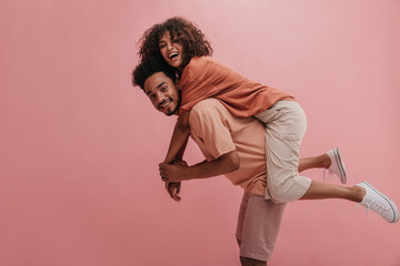 side profile full length cheerful positive cute couple in casual wear. african american man carrying woman smiling with teeth over isolated pastel pink background.