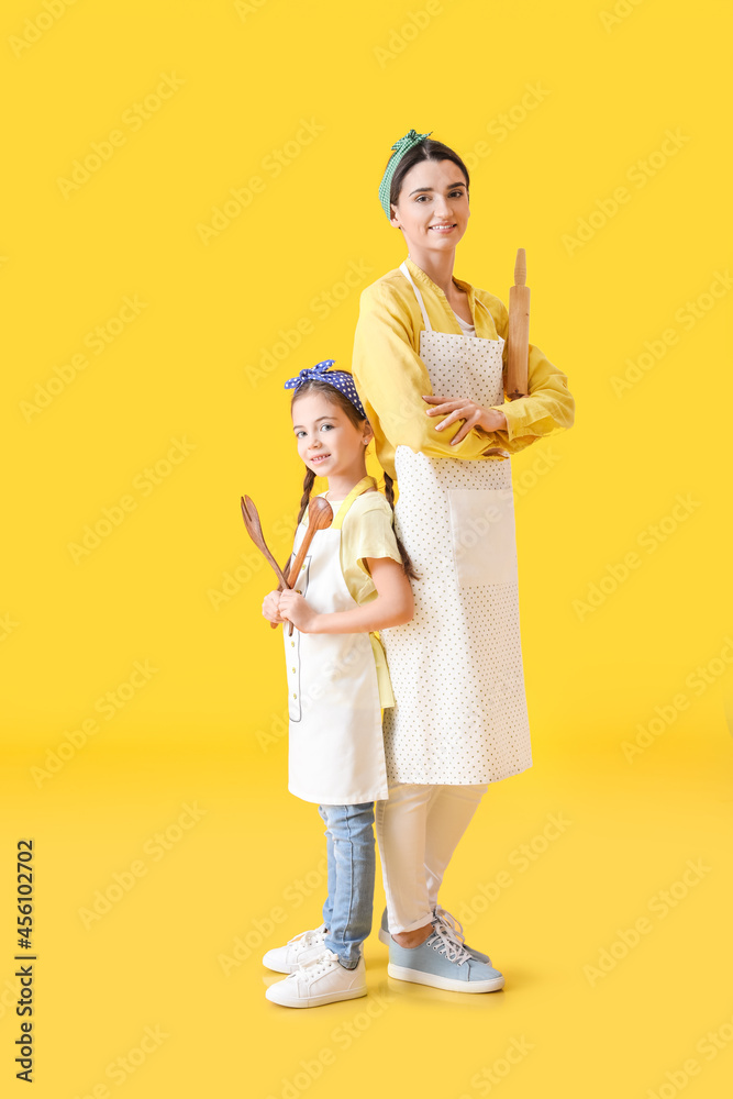 Young mother and daughter with kitchen utensils on color background