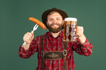 Wall Mural - Portrait of bearded man, waiter in traditional Bavarian costume with one liter of dark beer isolated over green background. Oktoberfest, festival, traditions concept