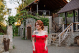 Fototapeta Londyn - Portrait of attractive redhead tattooed woman in red dress and diadema on blurred medieval castle background