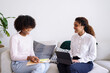 © sashapritchard - Two young business professional black women having interview conversation wearing smart casual sitting on sofa using tablet