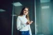 © BullRun - Half length portrait of successful female employee with digital cellular technology smiling at camera during work day in modern office interior, happy Caucasian entrepreneur with mobile phone