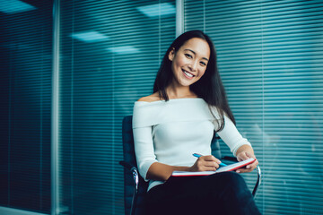 Portrait of Asian female employee with textbook creating business management and organization plan, cheerful professional woman enjoying own occupation holding notepad and smiling at camera