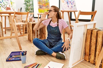 Canvas Print - Young brunette woman at art studio sitting on the floor shouting and screaming loud to side with hand on mouth. communication concept.