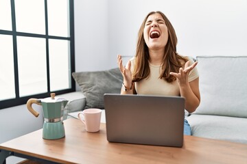 Poster - Young brunette woman using laptop at home drinking a cup of coffee crazy and mad shouting and yelling with aggressive expression and arms raised. frustration concept.