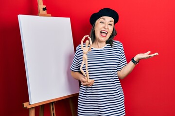 Wall Mural - Middle age hispanic woman standing by painter easel stand holding manikin celebrating achievement with happy smile and winner expression with raised hand