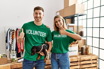 Sticker - Young couple with cute dog wearing volunteer t shirt at donations stand smiling happy and positive, thumb up doing excellent and approval sign