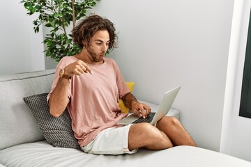 Poster - Young hispanic man sitting on the sofa at home using laptop pointing down with fingers showing advertisement, surprised face and open mouth