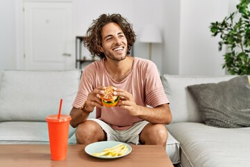 Poster - Young hispanic man smiling happy eating classic burger and drinking soda at home.