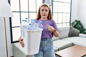 Sticker - Young redhead woman holding recycling wastebasket with plastic bottles pointing aside worried and nervous with forefinger, concerned and surprised expression