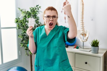 Poster - Young redhead physiotherapist woman holding prescription pills annoyed and frustrated shouting with anger, yelling crazy with anger and hand raised