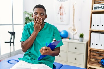 Sticker - Young african american man working at pain recovery clinic laughing and embarrassed giggle covering mouth with hands, gossip and scandal concept