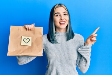 Wall Mural - Young modern girl holding take away paper bag with heart note smiling happy pointing with hand and finger to the side