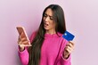 Young hispanic girl holding smartphone and credit card in shock face, looking skeptical and sarcastic, surprised with open mouth