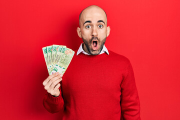 Sticker - Young bald man holding argentine pesos banknotes scared and amazed with open mouth for surprise, disbelief face