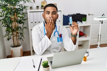 Sticker - Young indian doctor holding medical asthma inhaler at the clinic covering mouth with hand, shocked and afraid for mistake. surprised expression