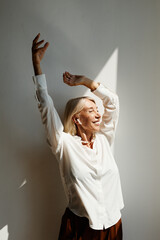 Wall Mural - Vertical portrait of elegant mature woman dancing lit by sunlight against white wall, copy space