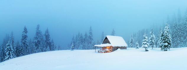 Wall Mural - fantastic winter landscape panorama with glowing wooden cabin in snowy forest. cozy house in carpath