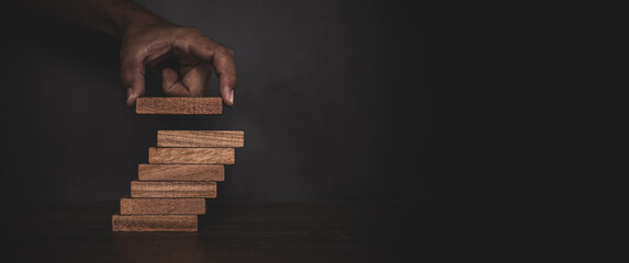 Close-up hand is placing wood block tower stacked in stair step with caution to prevent collapse or crash concepts of financial risk management and strategic planning.