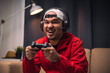© Chanakon - Playing video game. Young asian handsome man sitting on sofa holding joystick in living room. Happiness Streamer Indian man wearing headset playing game online in the darkroom.