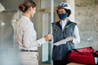 © Drazen - Food courier wears face mask while businesswoman signs for delivery in the office.