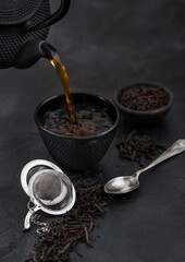 Pouring black tea from teapot to cup with strainer infuser and loose tea on black with silver spoon