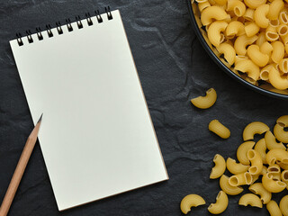 Wall Mural - open empty notebook and pencil with macaroni pasta on black background, copy space for text, food ba