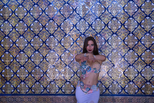 Middle-aged Hispanic woman in a beautifully colored dress with rhinestones to belly dance, making figures while dancing with a wall of beautiful tiles in the background. Belly dance concept.