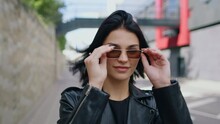 Portrait stylish seductive happy brunette woman wearing black leather jacket and sunglasses walking outdoors along city street looking at camera touches her hair flirts smiling. Urban style fashion