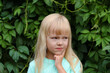 A little blonde girl in a mint T-shirt shows the emotion of resentment, sad. Brooding child against the background of green leaves. 