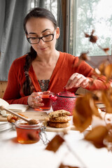 Woman wearing orange sweater pours a hot tea from red teapot in a glass mug on the table.Honey, biscuits and books near the cup of tea on the linen tablecloth.Fall vibes. Tea drinking. Selective focus