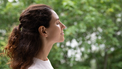 Profile close up face serene woman standing outdoor closed eyes breath fresh air feels motivated enjoy new day, green trees on background. No stress, filling with positive emotions, meditation concept