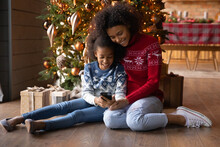 Happy Young African American Mother And Small Daughter Sit Near Fir Tree At Home Use Modern Cellphone Gadget. Smiling Biracial Mom And Girl Child Relax On Christmas Holiday Talk On Video Call On Cell.
