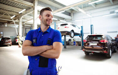 Wall Mural - Confident handsome young and experienced car repair worker in work overalls posing against the background of lifted cars in a car service