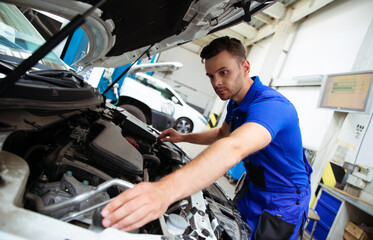 Wall Mural - Cheerful, handsome, and confident car repair specialist in overalls repairs and replaces old parts with new ones in a car on a lift in service