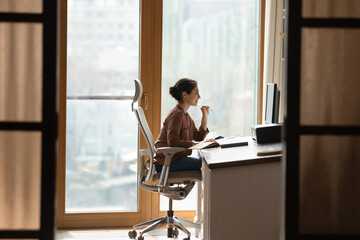 Working at modern home office. Young mixed race female architect interior designer sit at desk on ergonomic chair doing job project on desktop pc. Millennial indian woman study at domestic workplace