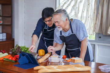 happy asian young son and senior old father cooking online class on laptop computer together making fresh vegetables food in kitchen at home