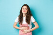 © Mix and Match Studio - Image of sad young woman frowning, holding hands on stomach, having ache, painful menstrual cramps, standing against blue background