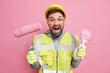 © WHstudio Leushin N - Positive male professional handyman holds painting roller and brush looks gladfully at camera happy to finish doing repairing dressed in uniform isolated over pink background. Maintenance work