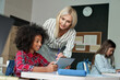 © insta_photos - Happy smiling Caucasian teacher explaining task to African American schoolgirl using tablet device sitting in classroom with group of schoolchildren. Modern technologies for education concept.