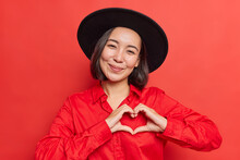 Gentle Smiling Asian Woman Shows Heart Sign Near Chest I Love You Gesture Wears Black Hat And Shirt Poses Against Vivid Red Background Wishes Happy Valentines Day. Romantic Message Compassion