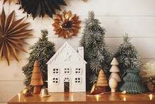 Stylish Christmas Little House, Trees, Golden Lights On Background Of Paper Stars On White Wooden Wall. Atmospheric Festive  Decor In Modern Scandinavian Room. Magic Winter Time. Merry Christmas!
