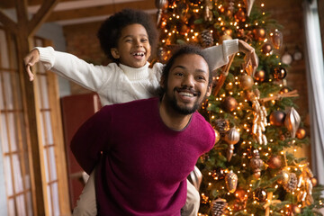 Wall Mural - Overjoyed small biracial boy child piggyback smiling African American father play together on Christmas holidays at home. Happy ethnic dad and son have fun near fir tree relax on New Year vacation.