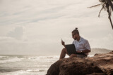 Fototapeta  - Cyfrowy nomada, człowiek pracujący z laptopem i smartfonem zdalnie na tle oceanu i skał siedzący na wybrzeżu.