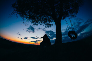 Silhouette of a tree with a tire swing on it at sunset and a photographer seating.