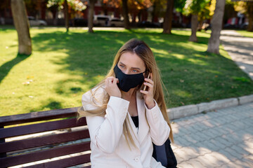 Wall Mural - Young woman wearing a medical protective mask sits on a park bench during the coronavirus pandemic and talks on a mobile video