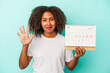 © Asier - Young african american woman holding a calendar isolated on blue background smiling cheerful showing number five with fingers.