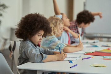 Wall Mural - Diligent little schoolboy writing in his notebook while studying, sitting at the table in elementary school classroom