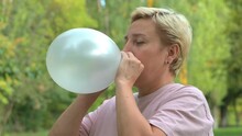 A Blonde Girl With Short Hair Inflates A Balloon Against The Background Of Green Nature Close-up