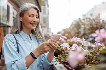 Wall Mural - Smiling grey haired mature Asian woman in stylish shirt uses cellphone standing on outdoors terrace decorated with pink flowers